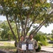Two U.S. Army soldiers in OCP (Operational Camouflage Pattern) uniforms are posing outdoors while holding framed certificates with blue borders. Both individuals are smiling, suggesting a celebratory or recognition occasion. The setting appears to be a military installation with trees, landscaping, and paved walkways visible in the background.