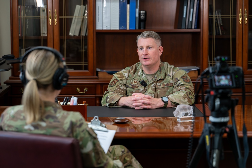 Two military personnel in OCP (Operational Camouflage Pattern) uniforms are engaged in a recorded interview setting. A male officer sits across a desk from a female soldier who is wearing headphones and holding a clipboard, while a camera on a tripod records the session. The setting appears to be a formal military office with wooden bookshelves in the background.