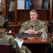 Two military personnel in OCP (Operational Camouflage Pattern) uniforms are engaged in a recorded interview setting. A male officer sits across a desk from a female soldier who is wearing headphones and holding a clipboard, while a camera on a tripod records the session. The setting appears to be a formal military office with wooden bookshelves in the background.