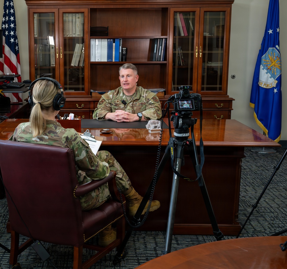 A military interview is being conducted in a formal office setting, with two uniformed personnel — one seated behind a large executive desk and one facing them with a clipboard and headphones. A Nikon camera on a tripod is positioned to record the interview. The setting includes U.S. Air Force and American flags, suggesting a senior officer's office.