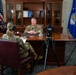 A military interview is being conducted in a formal office setting, with two uniformed personnel — one seated behind a large executive desk and one facing them with a clipboard and headphones. A Nikon camera on a tripod is positioned to record the interview. The setting includes U.S. Air Force and American flags, suggesting a senior officer's office.
