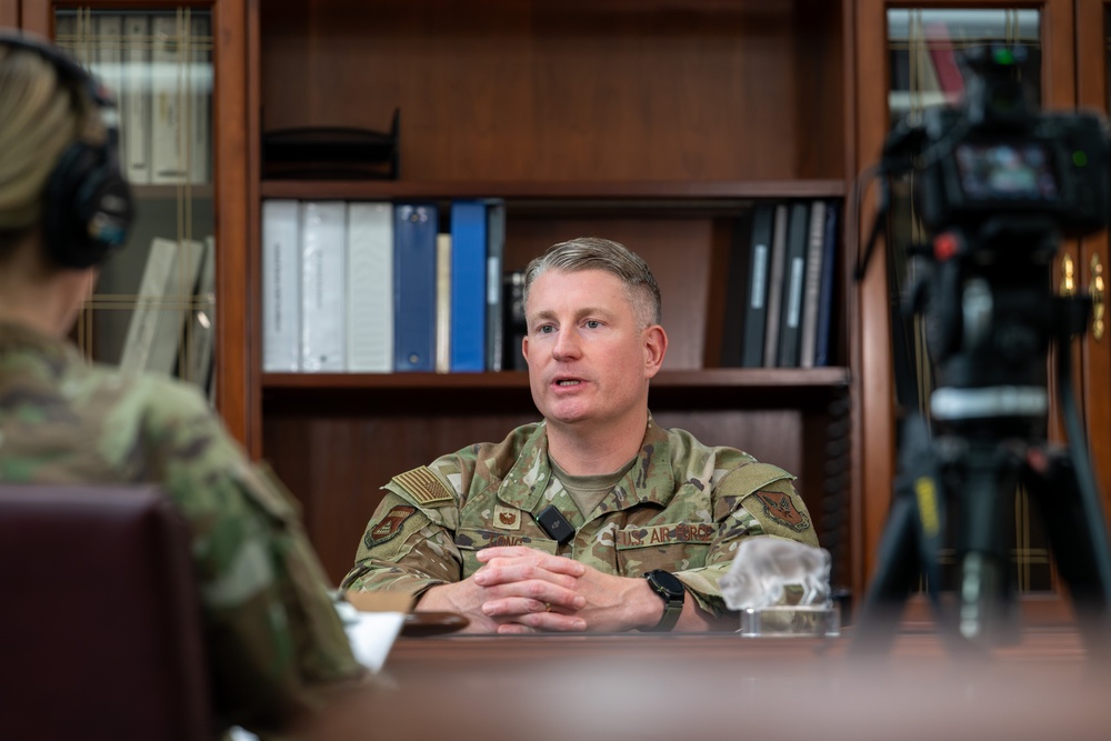 A U.S. Air Force officer, identifiable by the 'U.S. AIR FORCE' tape and OCP (Operational Camouflage Pattern) uniform, is seated at a desk being interviewed or recorded. The setting appears to be an office with a wooden bookshelf filled with binders and books in the background. A camera on a tripod and another uniformed individual wearing headphones are visible in the foreground.