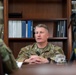 A U.S. Air Force officer, identifiable by the 'U.S. AIR FORCE' tape and OCP (Operational Camouflage Pattern) uniform, is seated at a desk being interviewed or recorded. The setting appears to be an office with a wooden bookshelf filled with binders and books in the background. A camera on a tripod and another uniformed individual wearing headphones are visible in the foreground.