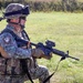 Two soldiers in camouflage uniforms are conducting what appears to be a military training exercise in an open grassy field. The foreground soldier is in a kneeling position holding an assault rifle with optical attachments, wearing full tactical gear including helmet and body armor. Both soldiers are equipped with weapons and protective equipment consistent with a live-fire or field training exercise.