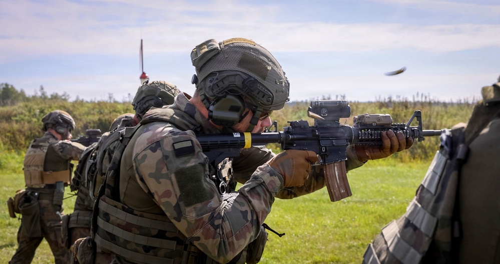 Military personnel are engaged in a live-fire training exercise on an open grassy range. The foreground soldier is actively firing a rifle, with a spent brass casing visibly ejecting from the weapon. Additional soldiers are visible in the background, also in firing positions.