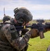 Military personnel are engaged in a live-fire training exercise on an open grassy range. The foreground soldier is actively firing a rifle, with a spent brass casing visibly ejecting from the weapon. Additional soldiers are visible in the background, also in firing positions.
