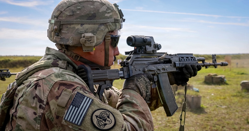 A U.S. Army soldier in MultiCam (OCP) uniform and helmet is aiming an AK-pattern rifle equipped with a modern optic at a firing range. The soldier's left shoulder displays a reversed U.S. flag patch and a unit patch. The setting appears to be an open outdoor range, likely during a familiarization or training exercise with foreign weapons.