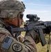A U.S. Army soldier in MultiCam (OCP) uniform and helmet is aiming an AK-pattern rifle equipped with a modern optic at a firing range. The soldier's left shoulder displays a reversed U.S. flag patch and a unit patch. The setting appears to be an open outdoor range, likely during a familiarization or training exercise with foreign weapons.