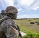 Military personnel in full combat gear are observed at what appears to be an outdoor firing range or tactical training area. The foreground soldier wears MultiCam/OCP camouflage, a combat helmet with a label, body armor, and carries a weapon. Shooting targets and hay bales are visible downrange, with a safety barrier tape marking the range boundary.