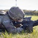 A soldier in full Operational Camouflage Pattern (OCP) uniform is in a prone firing position on a grass field, aiming down the sights of what appears to be an M4 carbine rifle equipped with an optic sight. The name tape on the helmet reads 'MUNES'. A second soldier is partially visible in the background in a similar position.