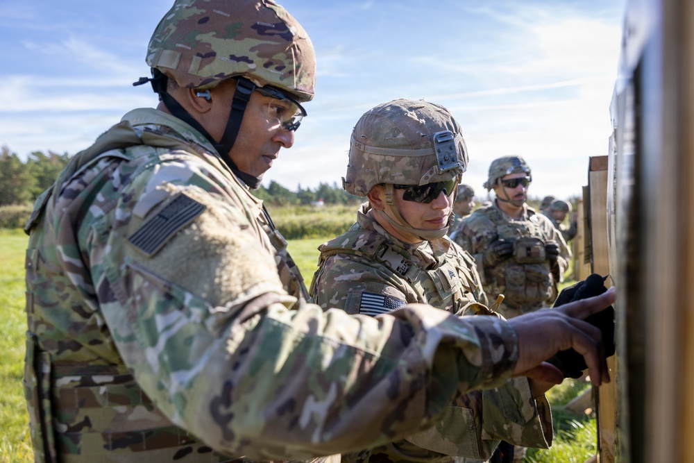 Two U.S. Army soldiers in the foreground are examining something on a board or panel, likely reviewing targets or instructions at a firing range or training exercise. Additional soldiers in full combat gear are visible in the background. The setting appears to be an outdoor military training area with open fields and trees.