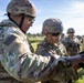 Two U.S. Army soldiers in the foreground are examining something on a board or panel, likely reviewing targets or instructions at a firing range or training exercise. Additional soldiers in full combat gear are visible in the background. The setting appears to be an outdoor military training area with open fields and trees.