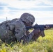 A soldier in full combat gear is in a prone firing position in an open grassy field, aiming an M16/M4-style rifle downrange. The soldier is wearing OCP (Operational Camouflage Pattern) uniform, tactical vest, and helmet. At least one additional soldier is partially visible in the background, suggesting a group training exercise.