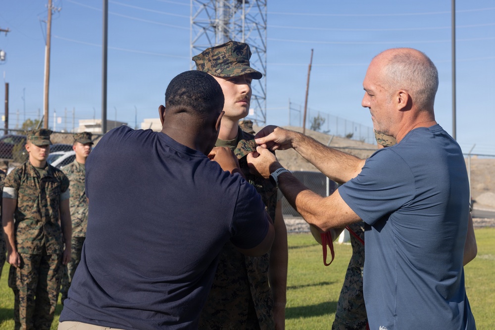 A military promotion or award ceremony is taking place outdoors on what appears to be a military installation. Two civilians in blue shirts are pinning something onto a uniformed Marine wearing MARPAT camouflage. Other Marines in uniform are visible standing in formation in the background.