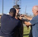 A military promotion or award ceremony is taking place outdoors on what appears to be a military installation. Two civilians in blue shirts are pinning something onto a uniformed Marine wearing MARPAT camouflage. Other Marines in uniform are visible standing in formation in the background.