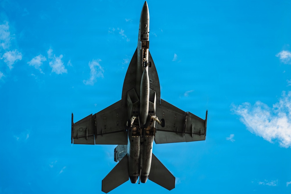 A military jet fighter is photographed from directly below, providing a ventral (bottom-up) view as it flies overhead against a bright blue sky with scattered clouds. The aircraft appears to be a twin-engine fighter jet, likely an F/A-18 Hornet or Super Hornet, based on its twin-tail configuration and overall planform shape. The aircraft is carrying external stores including what appear to be fuel tanks and possibly missiles on its hardpoints.