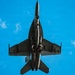 A military jet fighter is photographed from directly below, providing a ventral (bottom-up) view as it flies overhead against a bright blue sky with scattered clouds. The aircraft appears to be a twin-engine fighter jet, likely an F/A-18 Hornet or Super Hornet, based on its twin-tail configuration and overall planform shape. The aircraft is carrying external stores including what appear to be fuel tanks and possibly missiles on its hardpoints.