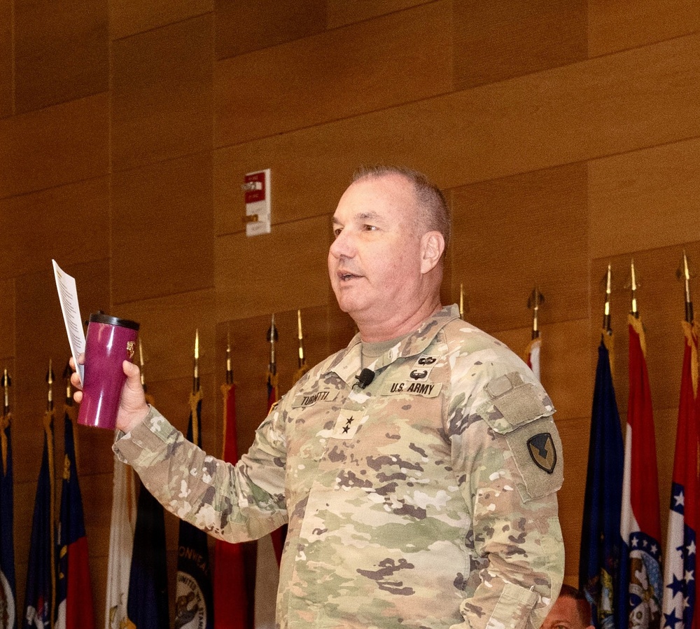 A U.S. Army Major General (two-star rank visible on collar) wearing OCP (Operational Camouflage Pattern) uniform is speaking at a formal military event or briefing. He is holding a purple/maroon tumbler cup and a sheet of paper, with a lapel microphone attached to his uniform. Multiple military unit guidons and state flags are displayed in the background.