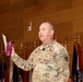A U.S. Army Major General (two-star rank visible on collar) wearing OCP (Operational Camouflage Pattern) uniform is speaking at a formal military event or briefing. He is holding a purple/maroon tumbler cup and a sheet of paper, with a lapel microphone attached to his uniform. Multiple military unit guidons and state flags are displayed in the background.