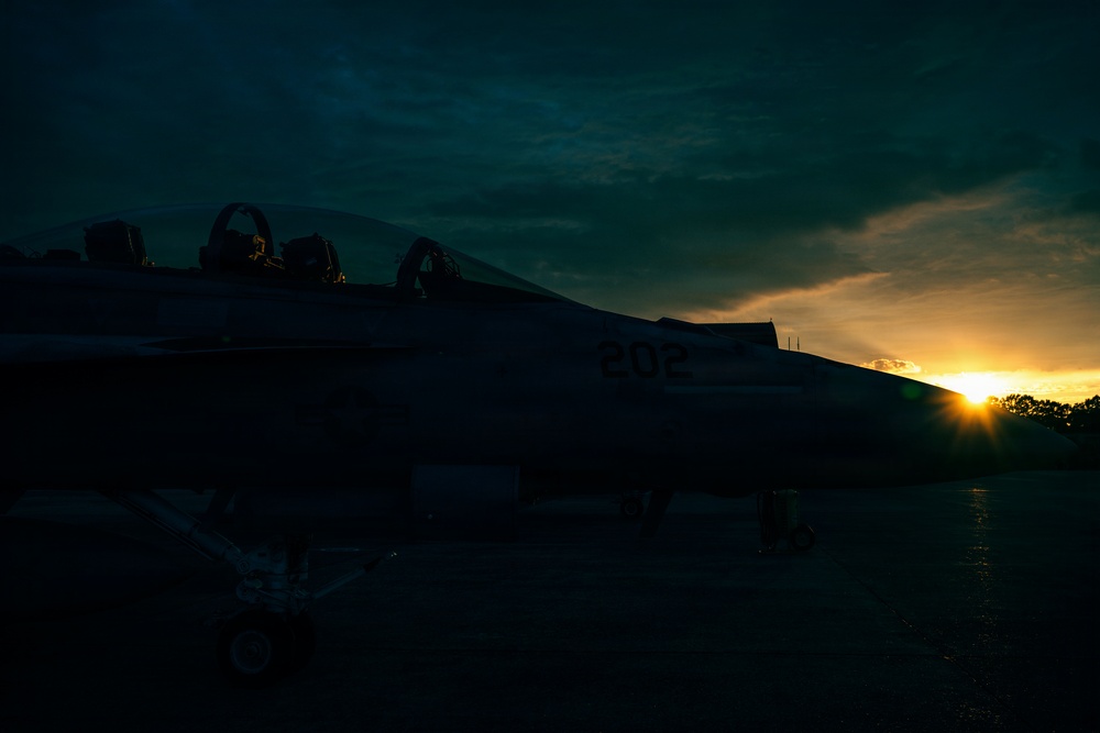 A military jet aircraft marked with the number '202' is photographed on a tarmac during sunset or sunrise. The aircraft is largely in silhouette against a dramatic sky with golden light breaking through clouds on the horizon. The two-seat cockpit with ejection seats is visible in the upper portion of the frame.