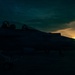 A military jet aircraft marked with the number '202' is photographed on a tarmac during sunset or sunrise. The aircraft is largely in silhouette against a dramatic sky with golden light breaking through clouds on the horizon. The two-seat cockpit with ejection seats is visible in the upper portion of the frame.