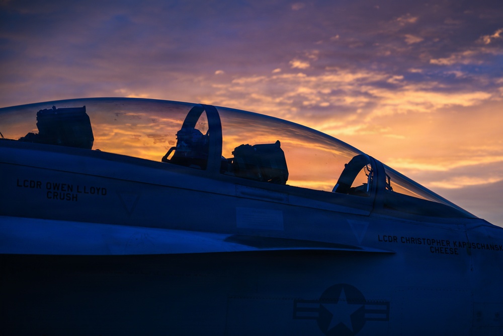 A U.S. Navy fighter jet, likely an F/A-18 Hornet or Super Hornet, is photographed at sunset or sunrise with dramatic orange and purple skies in the background. The cockpit canopy is open and pilot name stencils are visible on the fuselage. The image is a moody, silhouette-style photograph emphasizing the aircraft's profile against the colorful sky.