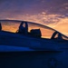 A U.S. Navy fighter jet, likely an F/A-18 Hornet or Super Hornet, is photographed at sunset or sunrise with dramatic orange and purple skies in the background. The cockpit canopy is open and pilot name stencils are visible on the fuselage. The image is a moody, silhouette-style photograph emphasizing the aircraft's profile against the colorful sky.