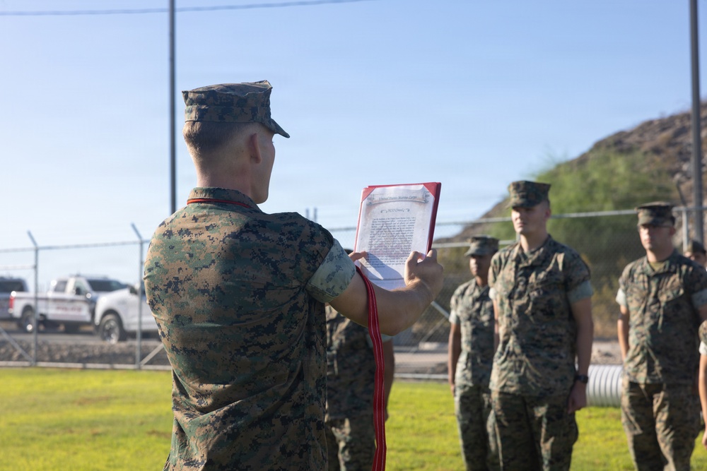 A U.S. Marine in digital camouflage (MARPAT) uniform is reading from a formal document held in a red folder to a formation of fellow Marines standing at attention outdoors. The document appears to be an official U.S. Marine Corps citation or commendation, identifiable by its scroll-style header. The ceremony is taking place in an outdoor military installation setting under clear skies.