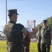 A U.S. Marine in digital camouflage (MARPAT) uniform is reading from a formal document held in a red folder to a formation of fellow Marines standing at attention outdoors. The document appears to be an official U.S. Marine Corps citation or commendation, identifiable by its scroll-style header. The ceremony is taking place in an outdoor military installation setting under clear skies.