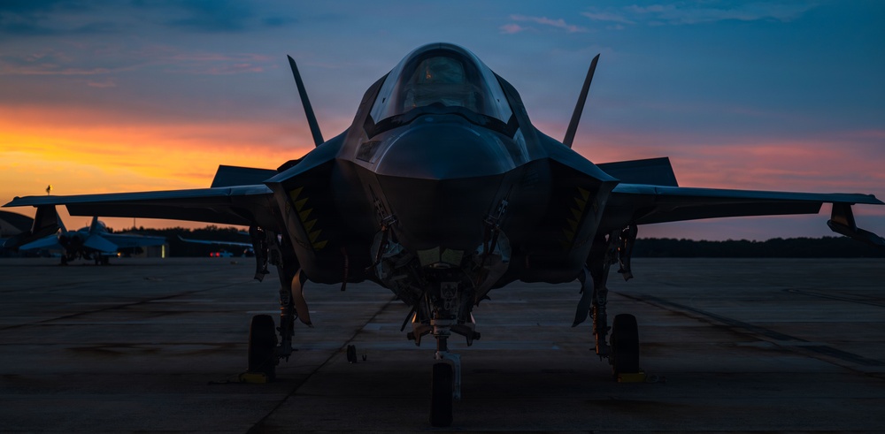 A military fighter jet, consistent with an F-35 Lightning II, is photographed head-on while parked on a flight line tarmac during twilight or sunset. The dramatic sky displays vivid orange, pink, and blue hues silhouetting the aircraft. At least one additional aircraft is visible in the background to the left.