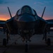 A military fighter jet, consistent with an F-35 Lightning II, is photographed head-on while parked on a flight line tarmac during twilight or sunset. The dramatic sky displays vivid orange, pink, and blue hues silhouetting the aircraft. At least one additional aircraft is visible in the background to the left.