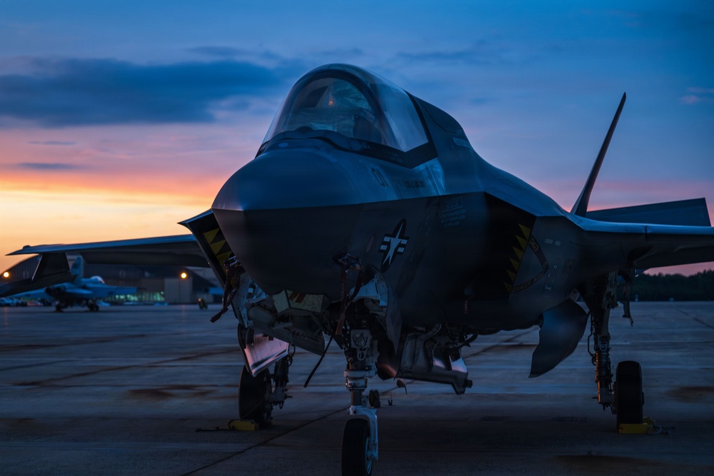 A fifth-generation stealth fighter jet, consistent with an F-35 Lightning II, is parked on a flight line apron during twilight or dawn. The aircraft is photographed from a front-facing low angle, with landing gear deployed and access panels open. A dramatic sky with orange, pink, and blue gradients provides a striking backdrop, with additional aircraft and hangar structures visible in the background.