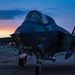 A fifth-generation stealth fighter jet, consistent with an F-35 Lightning II, is parked on a flight line apron during twilight or dawn. The aircraft is photographed from a front-facing low angle, with landing gear deployed and access panels open. A dramatic sky with orange, pink, and blue gradients provides a striking backdrop, with additional aircraft and hangar structures visible in the background.