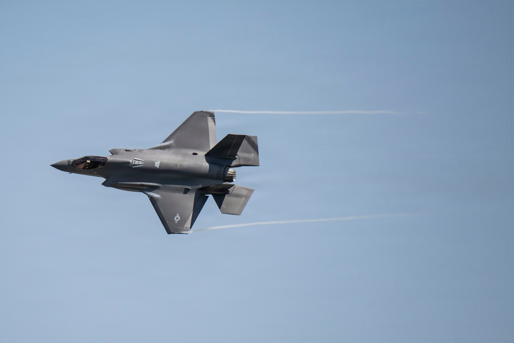 A single stealth fighter jet, consistent with an F-35 Lightning II, is captured in flight against a clear blue sky. The aircraft is viewed from a slightly elevated angle, showing its top surface with condensation trails streaming from its wingtips. The image is sharp and well-exposed, suggesting a professional aviation or airshow photograph.