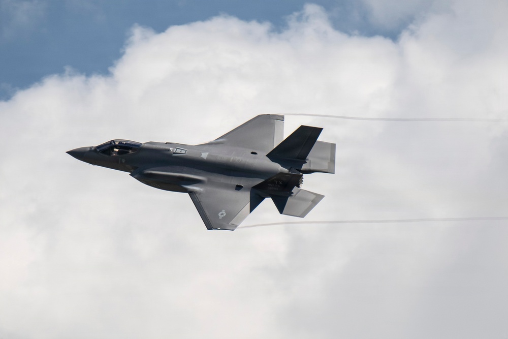 A single stealth fighter jet, consistent with an F-35 Lightning II, is captured in flight against a partly cloudy sky. The aircraft is banking slightly and generating visible wingtip vortex condensation trails. The image appears to have been taken at an airshow or during a demonstration flight.