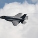 A single stealth fighter jet, consistent with an F-35 Lightning II, is captured in flight against a partly cloudy sky. The aircraft is banking slightly and generating visible wingtip vortex condensation trails. The image appears to have been taken at an airshow or during a demonstration flight.