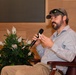 A bearded man wearing a camouflage cap with sunglasses resting on the brim is seated in a leather chair, speaking into a handheld microphone. He is dressed in a light blue checkered button-down shirt and khaki pants, appearing to be participating in a panel discussion or interview. The setting appears to be a formal venue with wood-paneled walls, a flag, and a decorative plant in the background.