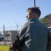 A person wearing a padded bite suit jacket and military camouflage pants is being bitten on the back by a dark-colored German Shepherd during what appears to be a military or law enforcement K9 training exercise. The dog is on a leash and actively biting the protective padding on the handler's back. The setting is an outdoor grassy area with a chain-link fence and utility structures visible in the background.