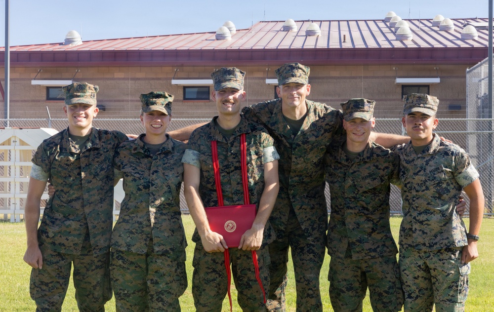 Six military personnel in MARPAT (Marine pattern) camouflage uniforms are posing together outdoors in a group photo. The central figure is holding a red-tasseled diploma or certificate folder bearing what appears to be a Marine Corps emblem, suggesting a graduation or completion ceremony. The group appears to be celebrating an academic or training milestone.