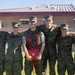 Six military personnel in MARPAT (Marine pattern) camouflage uniforms are posing together outdoors in a group photo. The central figure is holding a red-tasseled diploma or certificate folder bearing what appears to be a Marine Corps emblem, suggesting a graduation or completion ceremony. The group appears to be celebrating an academic or training milestone.