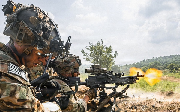 Two soldiers in full combat gear are firing a machine gun from a defensive fighting position or trench. The weapon is actively discharging, with a visible muzzle flash and numerous spent brass casings scattered on the ground. The setting appears to be a training exercise or live-fire drill in a rural, green landscape.