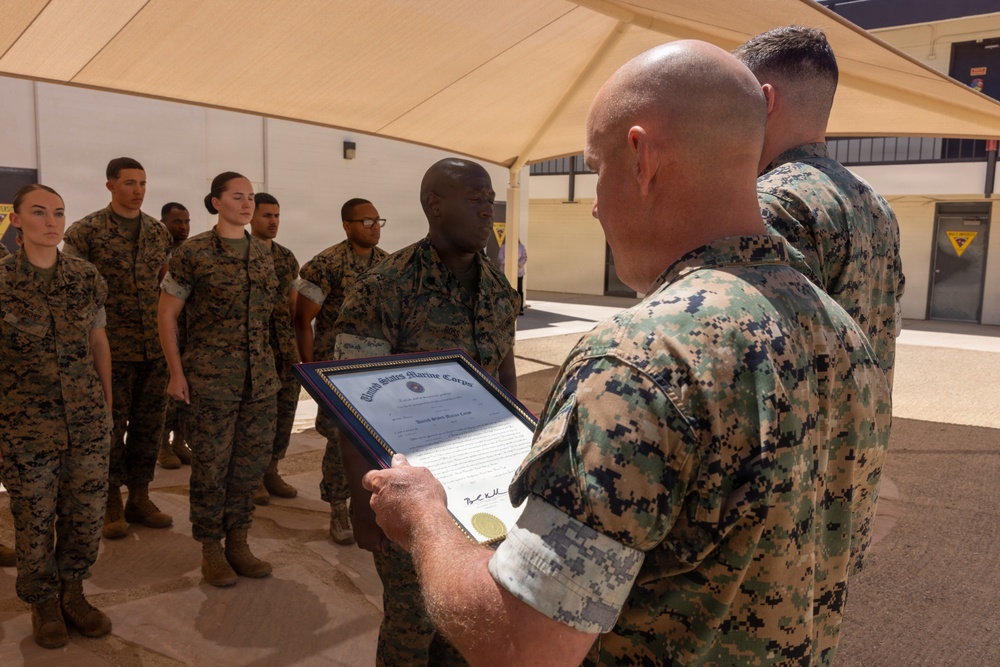 A United States Marine Corps ceremony is taking place outdoors under a tan shade canopy. A senior Marine is reading aloud from a framed official USMC certificate or commendation document to a recipient standing at attention. A formation of Marines in MARPAT camouflage uniforms stands in the background as witnesses.
