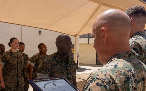 A United States Marine Corps ceremony is taking place outdoors under a tan shade canopy. A senior Marine is reading aloud from a framed official USMC certificate or commendation document to a recipient standing at attention. A formation of Marines in MARPAT camouflage uniforms stands in the background as witnesses.
