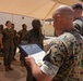 A United States Marine Corps ceremony is taking place outdoors under a tan shade canopy. A senior Marine is reading aloud from a framed official USMC certificate or commendation document to a recipient standing at attention. A formation of Marines in MARPAT camouflage uniforms stands in the background as witnesses.