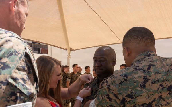 A military promotion or award ceremony is taking place outdoors under a shade canopy. A civilian woman in a floral dress and a uniformed Marine are pinning insignia or a medal onto a central Marine's uniform. Additional uniformed Marines are visible standing in formation in the background.