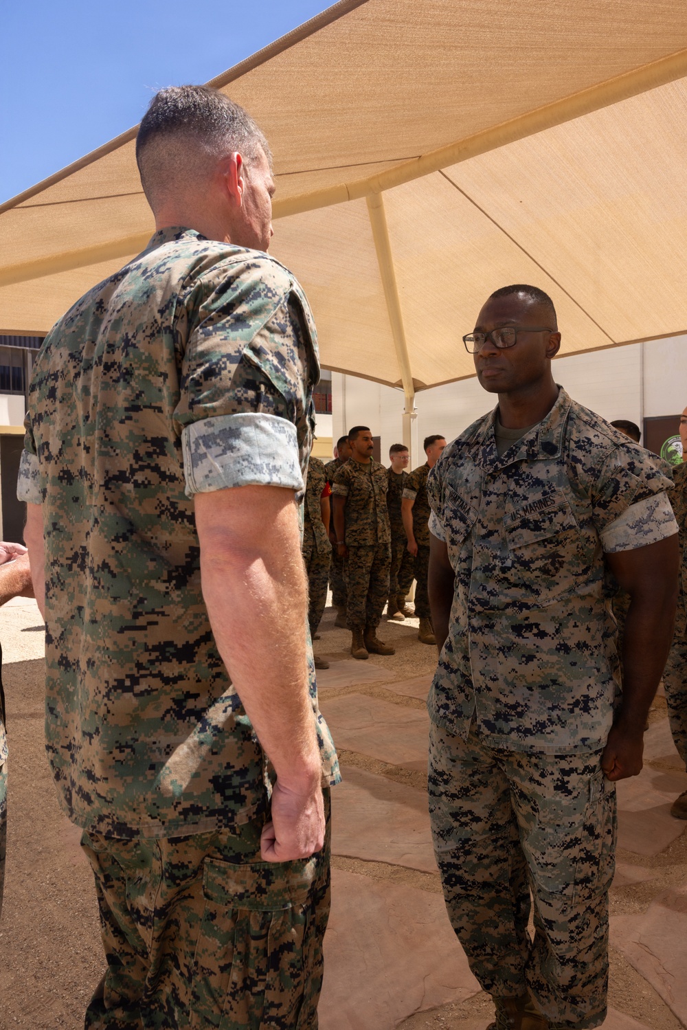 Two U.S. Marines in MARPAT digital camouflage uniforms are engaged in a face-to-face conversation under a tan shade canopy structure. The setting appears to be an outdoor military installation in an arid environment, with additional personnel visible in the background. The Marine facing the camera wears glasses and has 'U.S. MARINES' tape visible on his uniform.