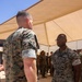 Two U.S. Marines in MARPAT digital camouflage uniforms are engaged in a face-to-face conversation under a tan shade canopy structure. The setting appears to be an outdoor military installation in an arid environment, with additional personnel visible in the background. The Marine facing the camera wears glasses and has 'U.S. MARINES' tape visible on his uniform.