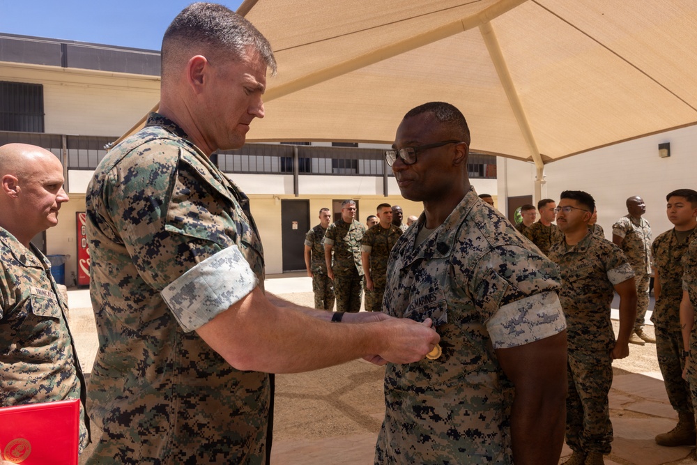 A military decoration or award ceremony is taking place outdoors at what appears to be a military installation. A senior Marine in MARPAT camouflage uniform is pinning a medal or award onto another Marine's uniform chest. A formation of Marines stands in the background observing the ceremony.