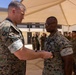 A military decoration or award ceremony is taking place outdoors at what appears to be a military installation. A senior Marine in MARPAT camouflage uniform is pinning a medal or award onto another Marine's uniform chest. A formation of Marines stands in the background observing the ceremony.