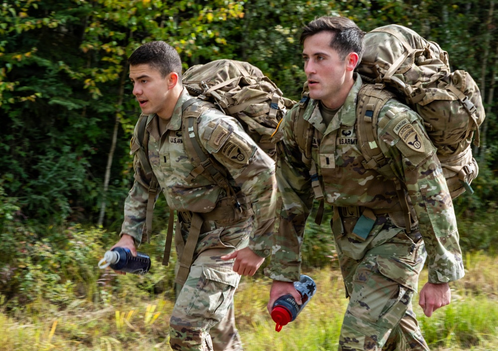 Two U.S. Army soldiers in Operational Camouflage Pattern (OCP) uniforms are conducting a ruck march through a wooded area. Both soldiers are carrying large military rucksacks and holding water bottles, suggesting an endurance training exercise. The soldier on the right displays a Ranger tab and what appears to be a 10th Mountain Division patch.